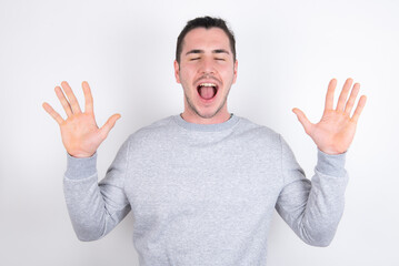 Naklejka premium Emotive Young handsome dark haired man wearing fitted T-shirt over white wall laughs loudly, hears funny joke or story, raises palms with satisfaction, being overjoyed amused by friend
