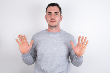 Serious Young handsome dark haired man wearing fitted T-shirt over white wall pulls palms towards...
