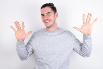 Fototapeta premium Young handsome dark haired man wearing fitted T-shirt over white wall showing and pointing up with fingers number ten while smiling confident and happy.