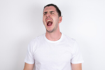 Young handsome dark haired man wearing fitted T-shirt over white wall angry and mad screaming...