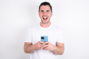 Excited Young handsome dark haired man wearing fitted T-shirt over white wall holding smartphone and looking amazed to the camera after receiving good news.
