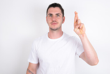 Fototapeta premium Young handsome dark haired man wearing fitted T-shirt over white wall pointing up with fingers number ten in Chinese sign language Shi