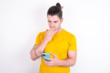 young dark haired man wearing yellow t-shirt over white background being deeply surprised, stares...