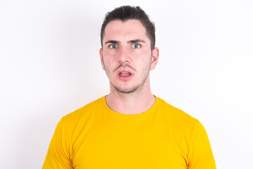 Obraz premium Shocked young dark haired man wearing yellow t-shirt over white background stares bugged eyes keeps mouth opened has surprised expression. Omg concept