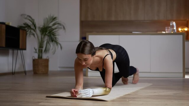 Young beautiful woman with bionic arm is doing plank exercise on floor in apartment room spbas. 4k Close view of caucasian female starts training and leans hands on mat, makes effort and poses for