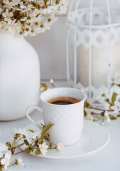 A beautiful postcard. A white coffee cup with a saucer, a statuette, candles and a vase with a bouquet of cherry blossoms. Beautiful still life. Spring time. The concept of "Good morning".