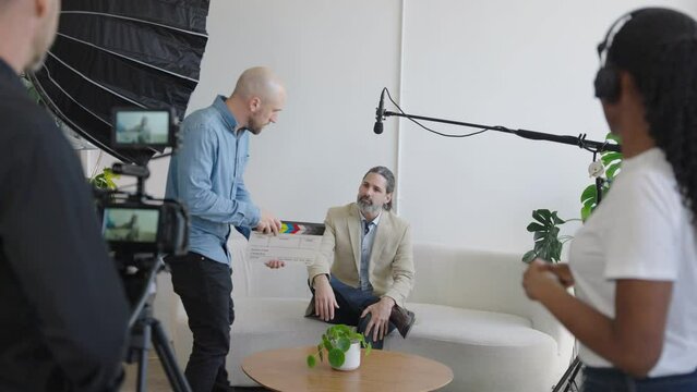 Behind the Scenes on a Video Production Set. Director Uses Clapper Slate to Signify Action. Camera Man, Audio Person, Boom Bole, Lighting and Video Camera Can Be Seen in Frame.