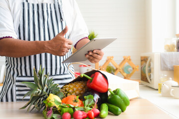 Close-up of chef's hand using tablet to buy fruit and vegetables online. And there are fruits and vegetables and paper bags on the kitchen table.shopping online and delivery concept.