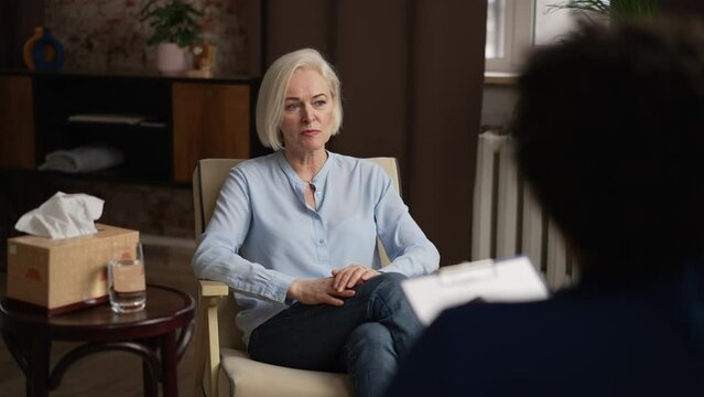 Elderly woman is talking to psychologist and sitting in chair in modern counseling office spbas. 4k Beautiful female patient shares problem and looks with smile, psychotherapist listens to her