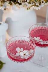 Two fashionable glasses for dessert and champagne with a pink drink and cherry blossoms inside on a light background with hard shadows and highlights. The concept of a party and a holiday.