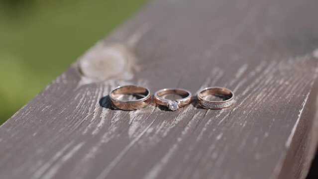 Three gold wedding and engagement rings of different sizes lie on painted and worn textured wooden plank of terrace railing. Mowed green lawn on background, cloudy.