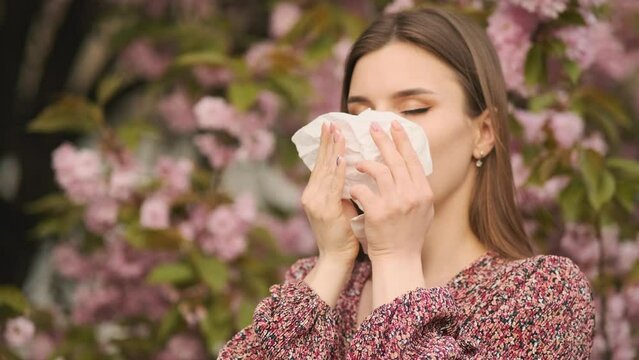 Spring allergy concept. Sneezing young girl with nose wiper among blooming trees in park. Pollen allergy, girl sneezing