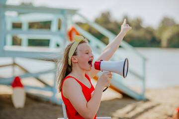 Pretty teen girl in red bikini posing with megaphone sitting on white summer chair at the beach...