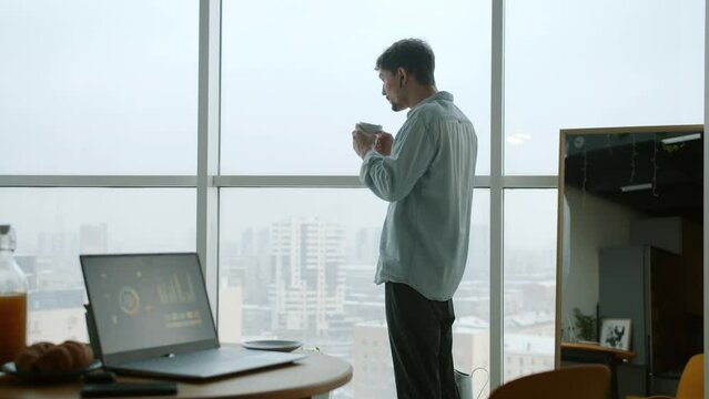Pensive young man in casual clothing is drinking coffee looking at city from apartment window. Laptop with business information is visible on table.