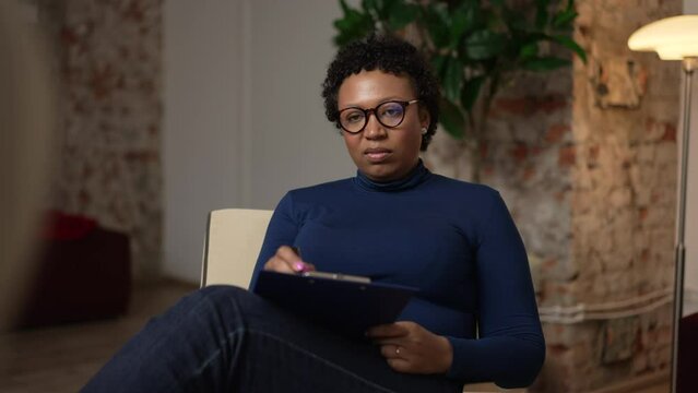 Portrait of young female psychoanalyst is making notes during psychotherapy session in office spbas. 4k American African woman listens to patient's story and shows empathy, writes on clipboard and