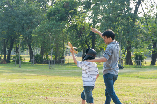 Happy Asian father playing paper plane with son at park, kid wearing pilot helmet, holding airplane toy in the air, Dad support to his dream goal, family insurance, copy space