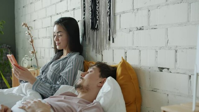 Happy Asian lady is having fun with smartphone while boyfriend is sleeping in bed at home. Modern devices and marital life concept.