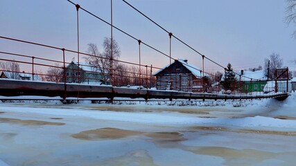 Suspension pedestrian bridge over a frozen river with wooden houses on the shore.