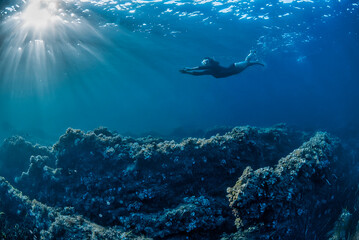 Girl swimming underwater in a rocky bottom with sunbeams coming from the left