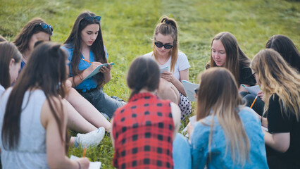 A group of female students are sitting in a circle on a meadow for collective work with notebooks.