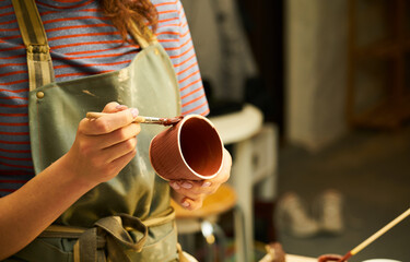 Close-up of girl painting clay mug with glaze. Woman coloring pottery in workshop with a paintbrush. Painter in green apron glazing clay pot.