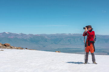 Fototapeta na wymiar Tourist in red walks on snow mountain near abyss edge on high altitude under blue sky in sunny day. Man with camera on snowy mountain near precipice edge with view to large mountain range in away.