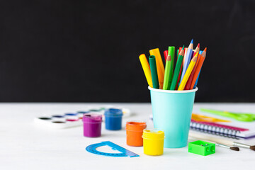 Obraz premium Time to return back to school. Bright colourful green yellow, blue stationery, paints, pencils, markers, brushes. black board on background. White alarm clock. Copy space for text. Autumn, September