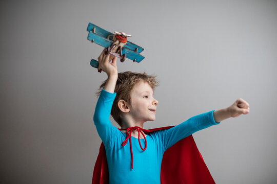 Boy dressed as a super hero in a red cape plays with an airplane at home. Travel.