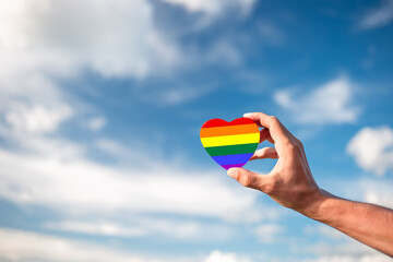 Hand holding painted rainbow heart symbolizing gay and lgbt pride and sky with clouds in the...