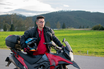 Obraz premium hombre parado al lado de una moto con un casco azul en la mano y fondo de montañas 