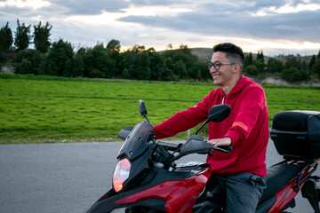 hombre sonriendo, con camisa roja, sentado en una moto con paisaje de arboles y cielo despejado 