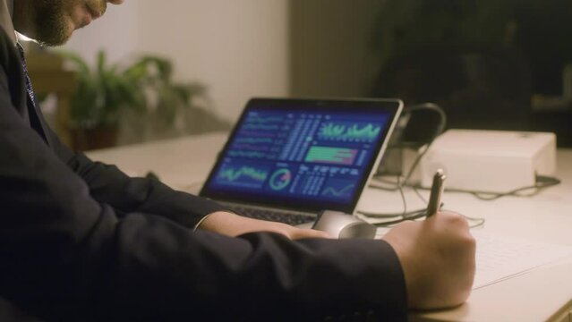 Caucasian bearded man working in office while typing on laptop and making notes. Serious man in suit analyzing charts on netbook screen while working alone. Business, work concept