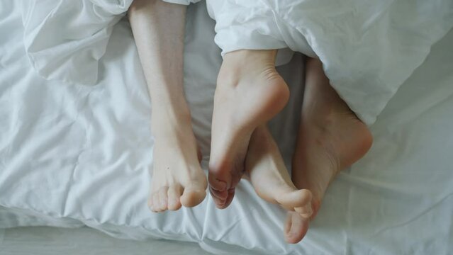 Close-up of male and female feet touching caressing expressing love in bed while couple man and woman lying in bedroom together. Relationship and intimacy concept.