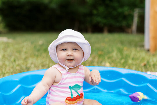 Adorable baby girl sits in a kiddy pool wearing a hat and swimsuit with chunky cheeks. 