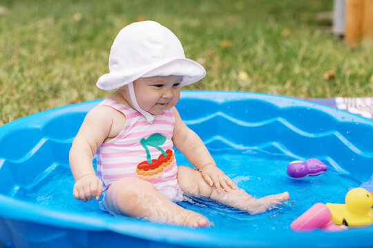 Adorable baby girl sits in a kiddy pool wearing a hat and swimsuit with chunky cheeks. 