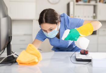 Woman in protective mask wipes dust from table in the office