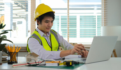 Professional Asian male architect using laptop computer, working on construction plan at his workplace.