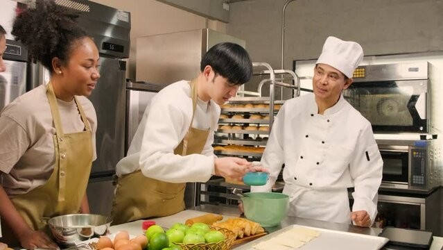 Cuisine course, senior male chef in uniform teaches young cooking class students, brushes pastry dough with eggs cream, prepares ingredients for bakery foods, fruit pies in stainless steel kitchen.
