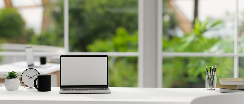 Modern white workspace tabletop with laptop mockup over blurred green office in the background.