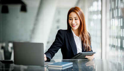 Photo of cheerful pretty Asian woman having been employed to a job as executive smiling toothily sitting at the desktop with laptop and writing note.