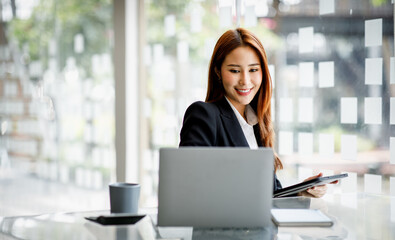 Photo of cheerful pretty Asian woman having been employed to a job as executive smiling toothily sitting at the desktop with laptop and writing note.