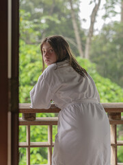 Back view of a Little Caucasian Girl relaxing on a wooden balcony in the forest with green background wearing a white bathrobe 