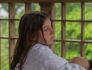 Little Caucasian Girl relaxing on a wooden balcony in the forest wearing a white bathrobe with a sad face. 