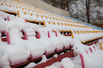 Empty outdoor football soccer stadium seats covered with snow in winter, light snowfall. Plastic seats in a row at the stadium are covered with snow snow.