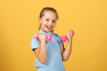 Cute little girl doing exercises with dumbbells in yellow background. Closeup portrait of sporty child with dumbbells. Happy child girl exercising.