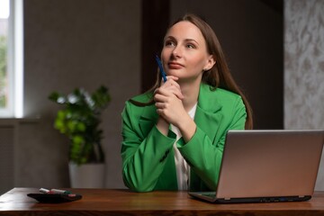 Pensive woman sits at desk near laptop holding pen in hand. Young brunette employee in green jacket dreams of vacation smiling at workplace in office