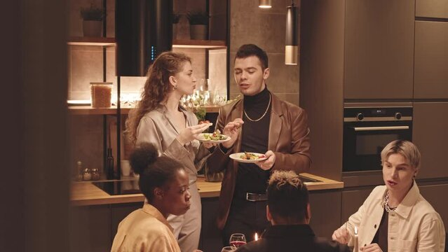 Medium slowmo of five fancy young LGBTQ friends eating and talking during small cozy dinner party in kitchen with dimmed light, having good time together