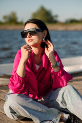 Stylish young woman dressed in casual in sunglasses sitting on wooden pier in sunny day