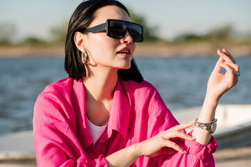 Stylish young woman dressed in casual in sunglasses sitting on wooden pier in sunny day