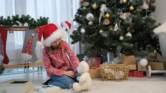Little girl in santa hat playing with teddy bear recieved as present at christmas sitting on floor under decorated tree